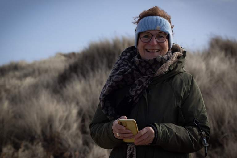Lächelnde Frau mit Handy, eingepackt in eine Jacke, vor plüschigen Dünen.
